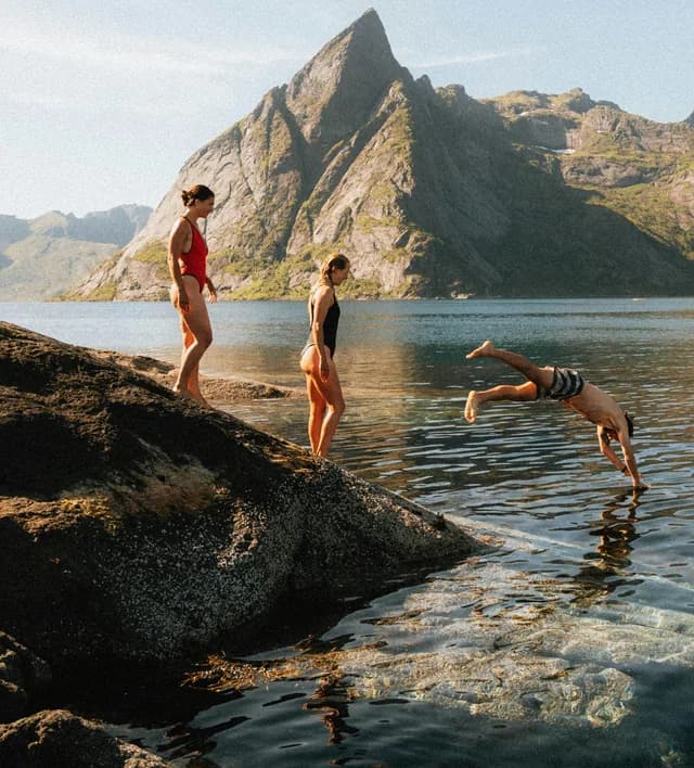 Three people swimming and diving off rocks in a fjord below a mountain peak