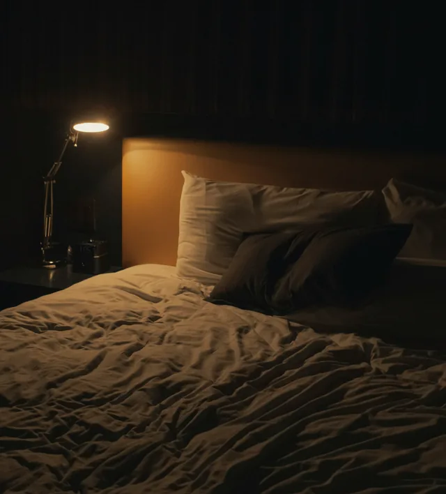 An unmade bed in a dim room lit by a single reading lamp